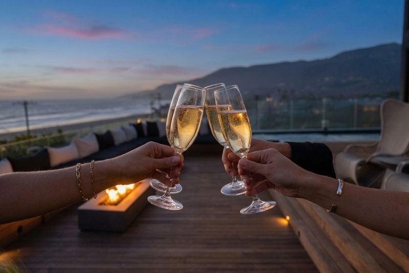 Four flutes of sparkling wine raised in a toast on an oceanfront deck at dusk, the fire table glowing behind them