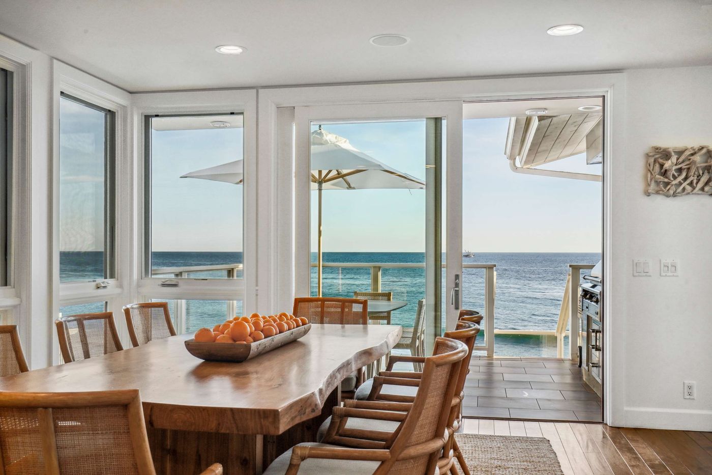 Dining table set beside windows and an open door with a deck and ocean view at an oceanfront Malibu home